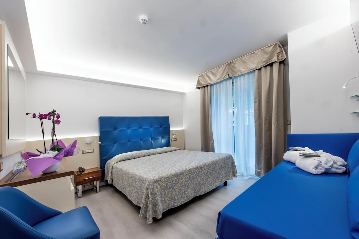 A hotel room with a bed, a desk with a chair, a small table with a vase of flowers, and a window with curtains.