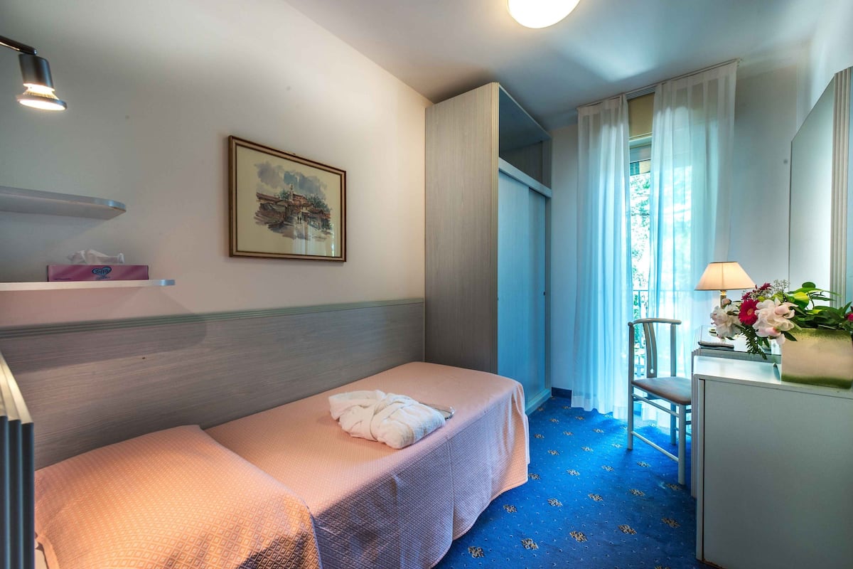 A hotel room with a single bed, a desk with a lamp, a chair, and a window with curtains.