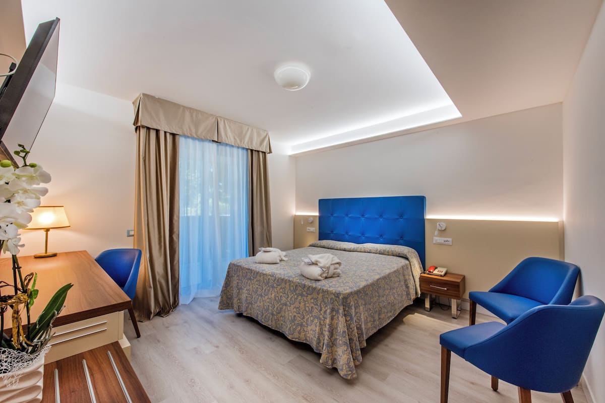 A modern hotel room with a bed, a desk, a chair, and a small table with a lamp.