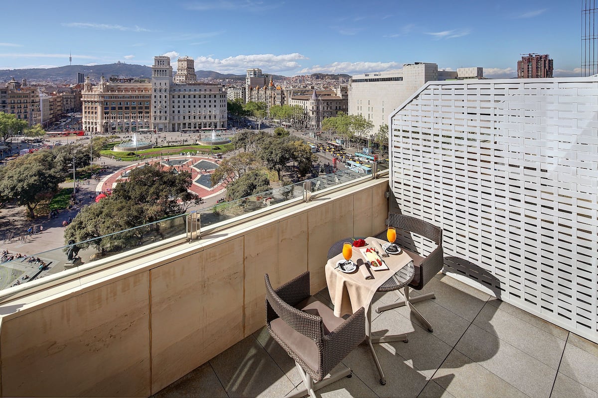 A balcony with a table set for two, overlooking a cityscape with historic buildings and a fountain.