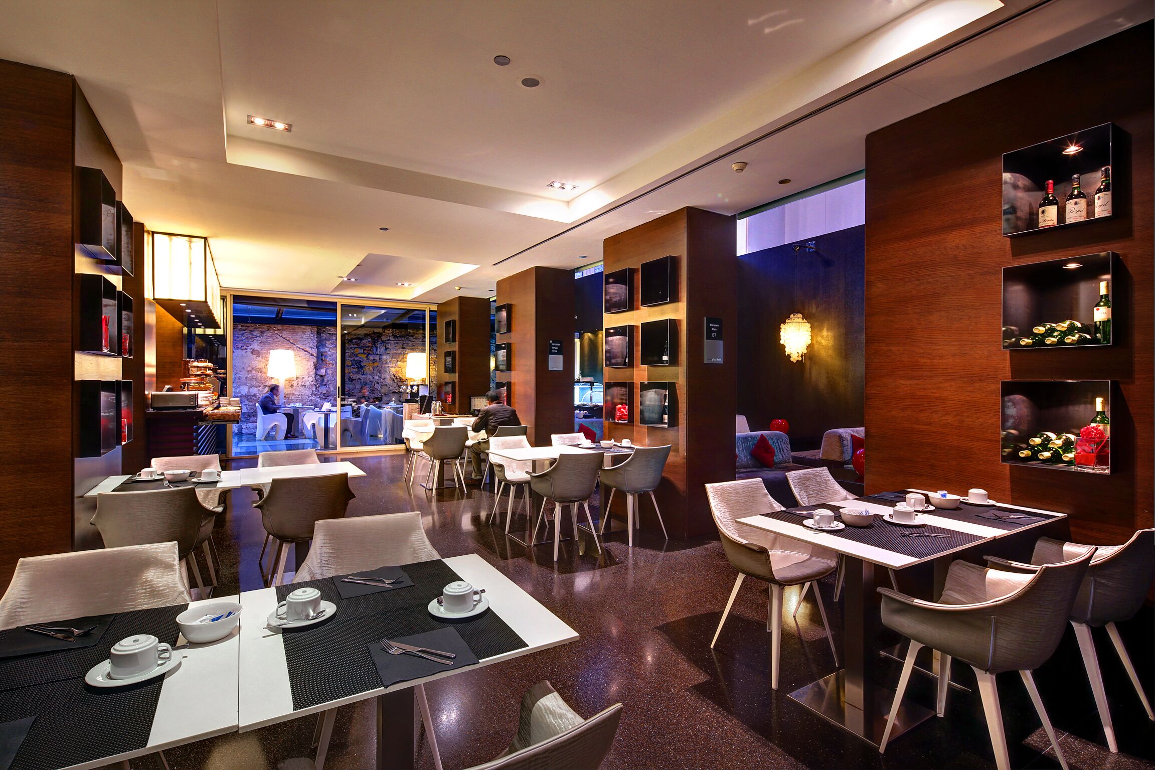 Un moderno vestíbulo de hotel con diferentes zonas de estar, incluyendo mesas y sillas, y un bar visible al fondo.
