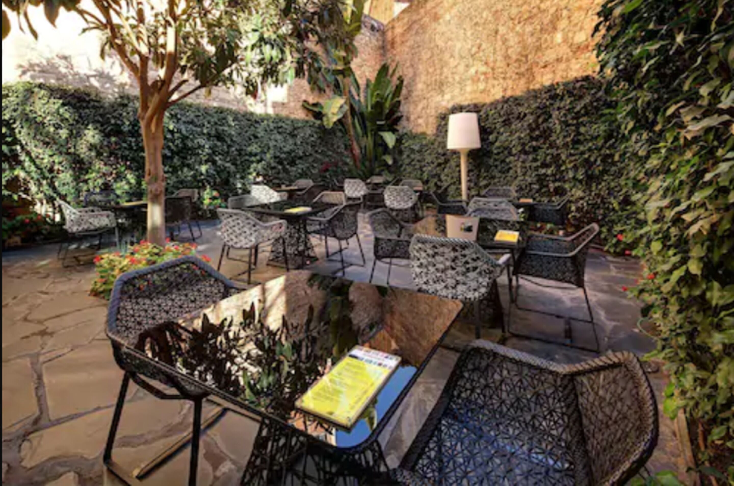 An outdoor seating area with wicker chairs and tables, surrounded by greenery and a stone wall.