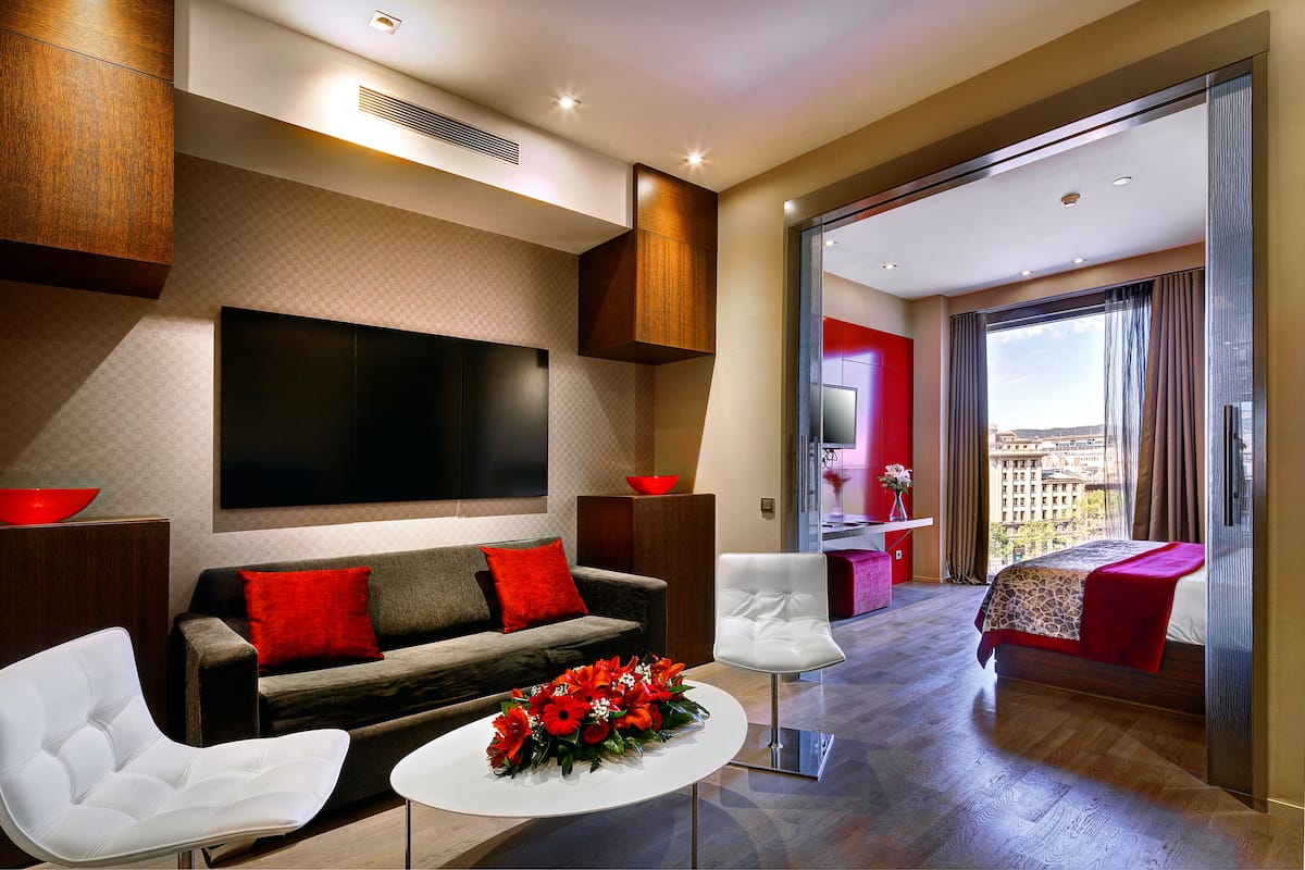 A modern hotel room with a large flat-screen TV, a sofa, a white armchair, a coffee table with a floral arrangement, and a balcony with a city view.