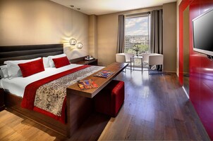 A hotel room with a large bed, a dining table, a TV, and a view of the city.