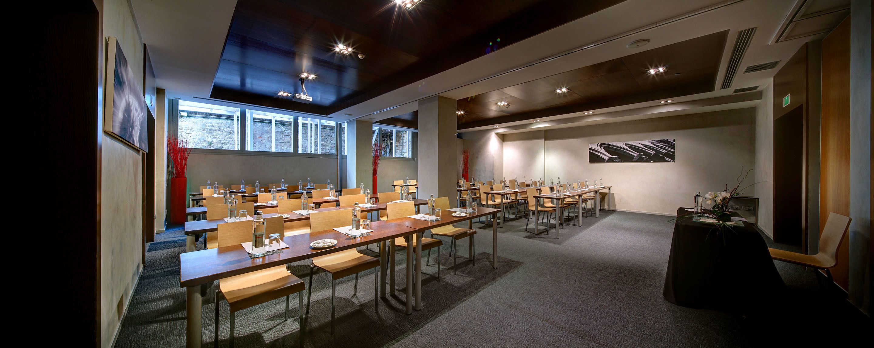Un salón de conferencias con varias mesas y sillas, ventanas amplias e iluminación en el techo.