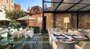 Un área de comedor al aire libre con mesas y sillas de vidrio, rodeada de muros de piedra y follaje.