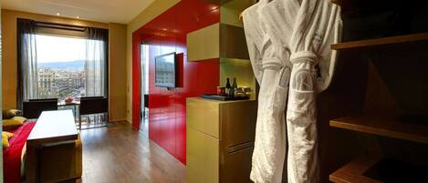 A hotel room with a dining area, a TV, a minibar, and a view of the city.