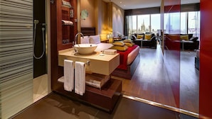 Een moderne hotelkamer met een groot bed, een zithoek met een bank en fauteuils, een groot raam met uitzicht op de stad, een badkamer met een grote wastafel en spiegel, en een houten vloer.