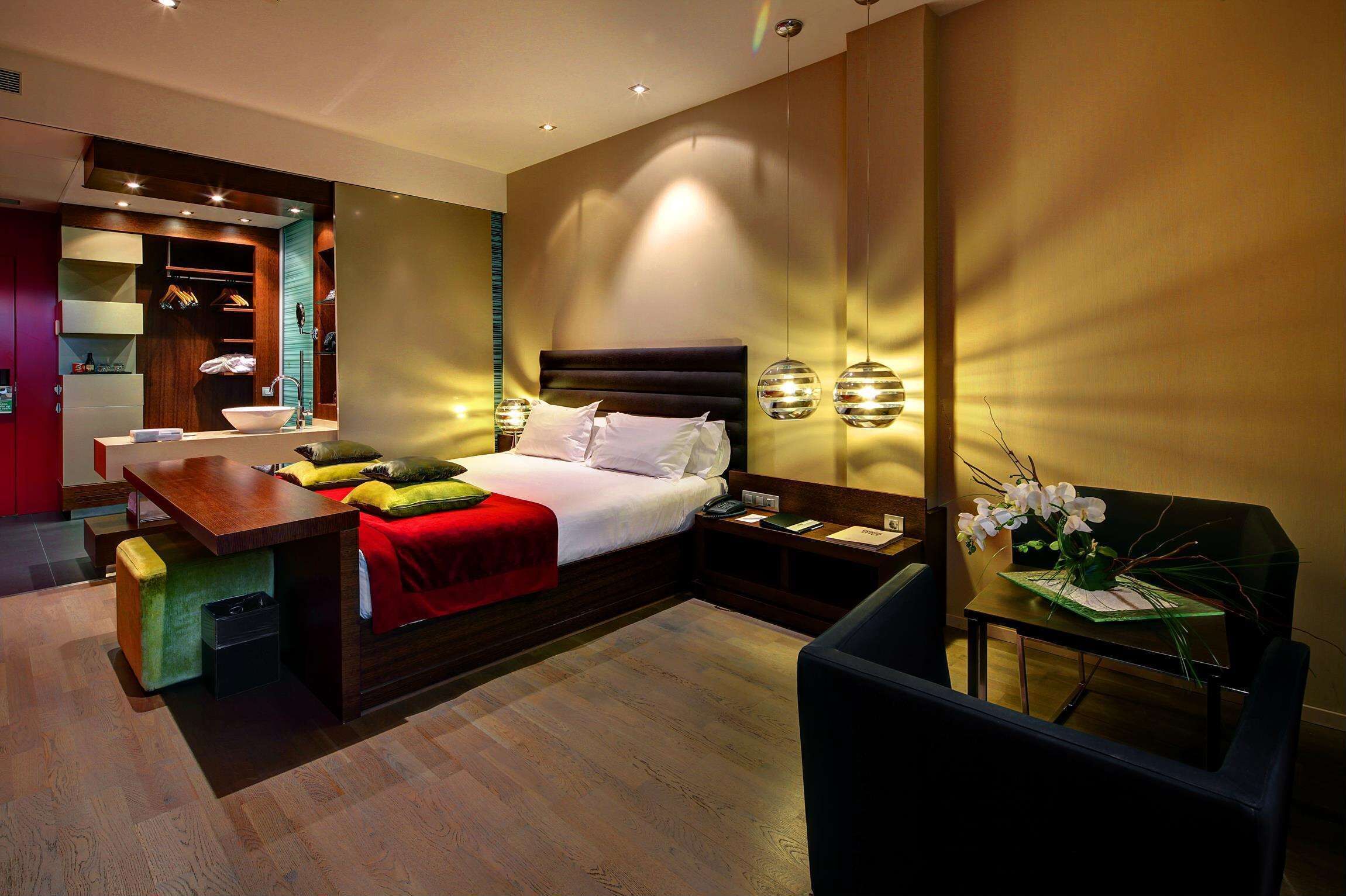 Ein modernes Hotelzimmer mit Bett, Nachttisch, Sessel und einem kleinen Tisch mit Blumenvase.