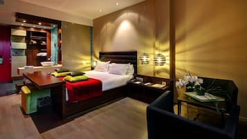 A modern hotel room with a bed, bedside table, chair, and a small table with a vase of flowers.