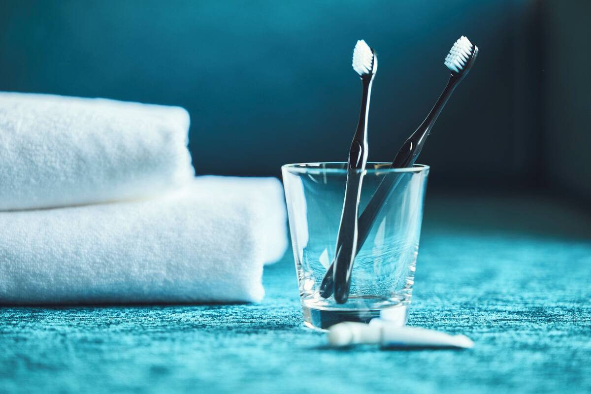 Un verre transparent contenant deux brosses à dents et un tube de dentifrice, posé sur une surface bleue texturée, avec une serviette blanche en arrière-plan.