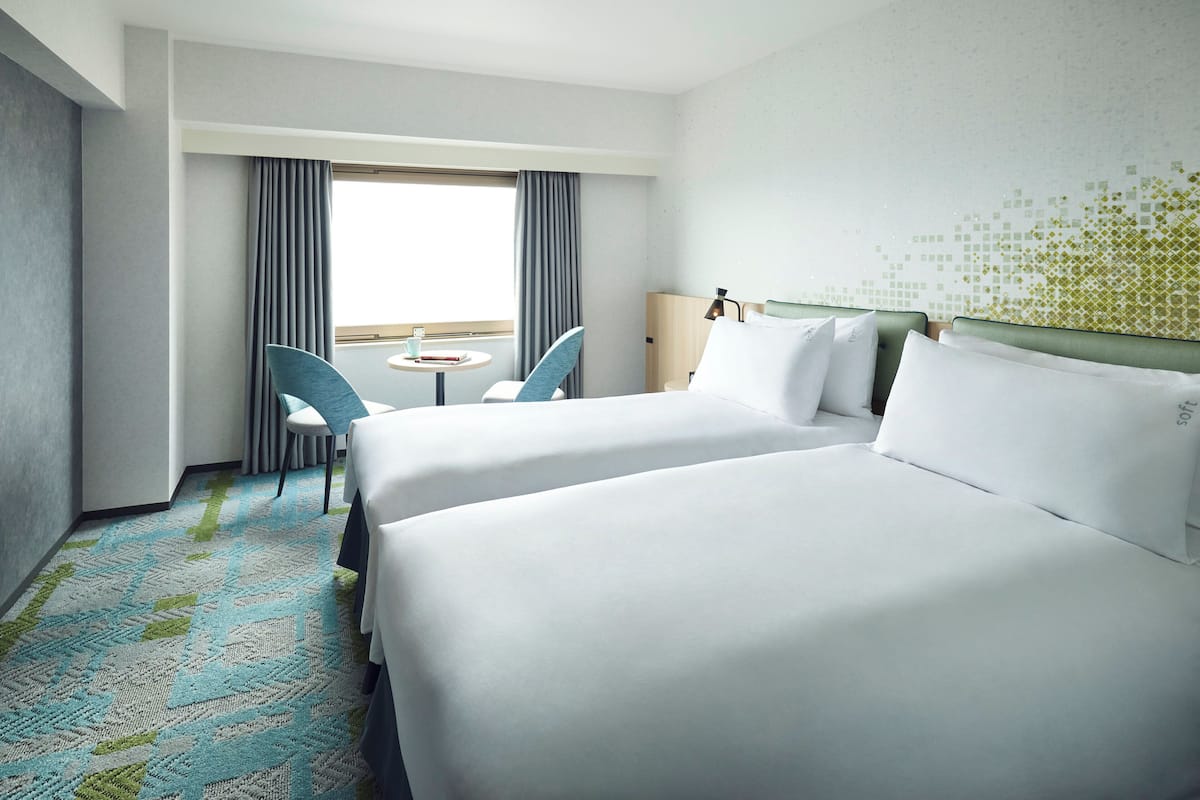 Une chambre d’hôtel avec deux lits, une petite table, deux chaises, une fenêtre avec des rideaux et une moquette à motifs.