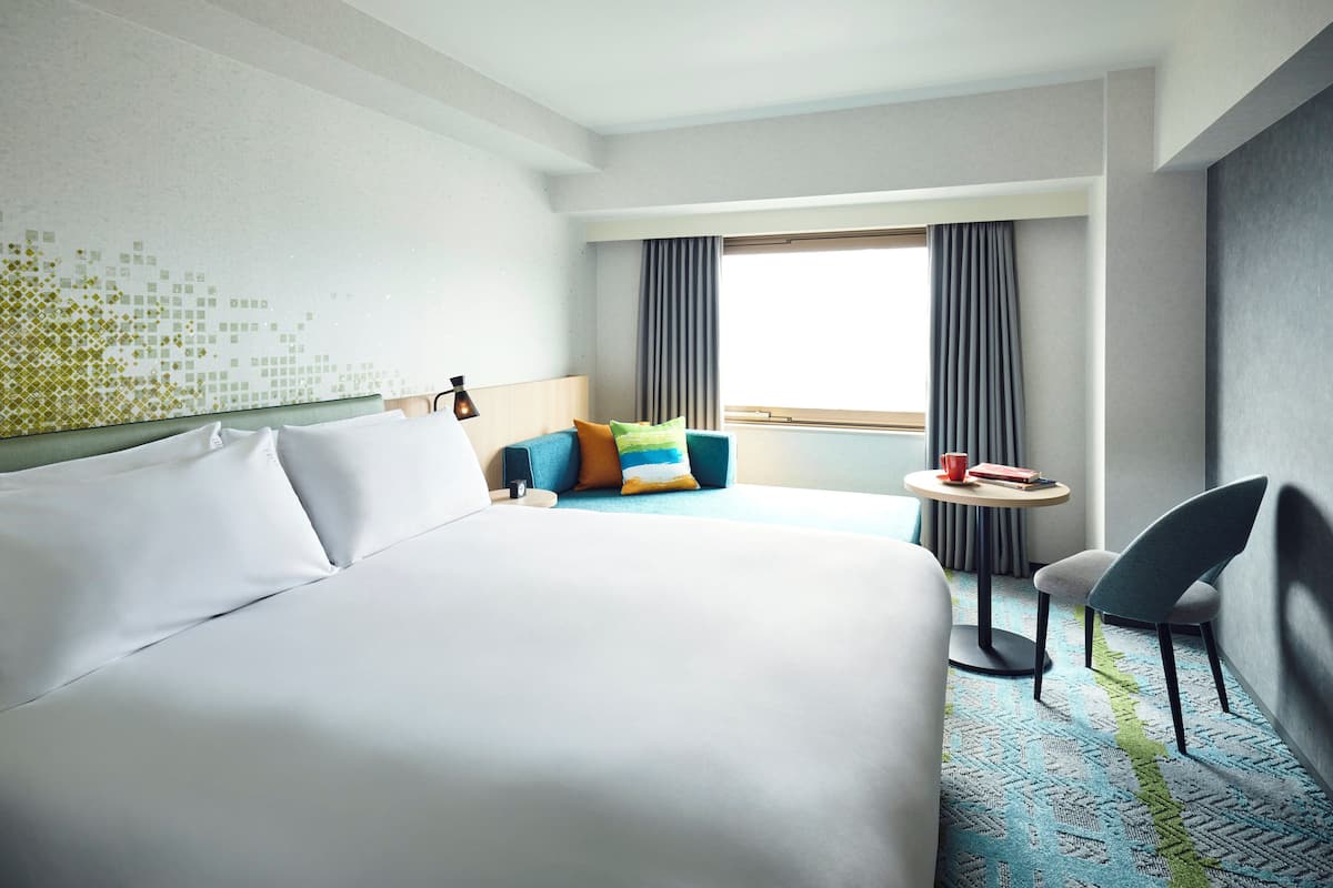 Une chambre d’hôtel avec un grand lit, une petite table avec une chaise, un canapé agrémenté de coussins colorés et une vue sur l’extérieur par une fenêtre.