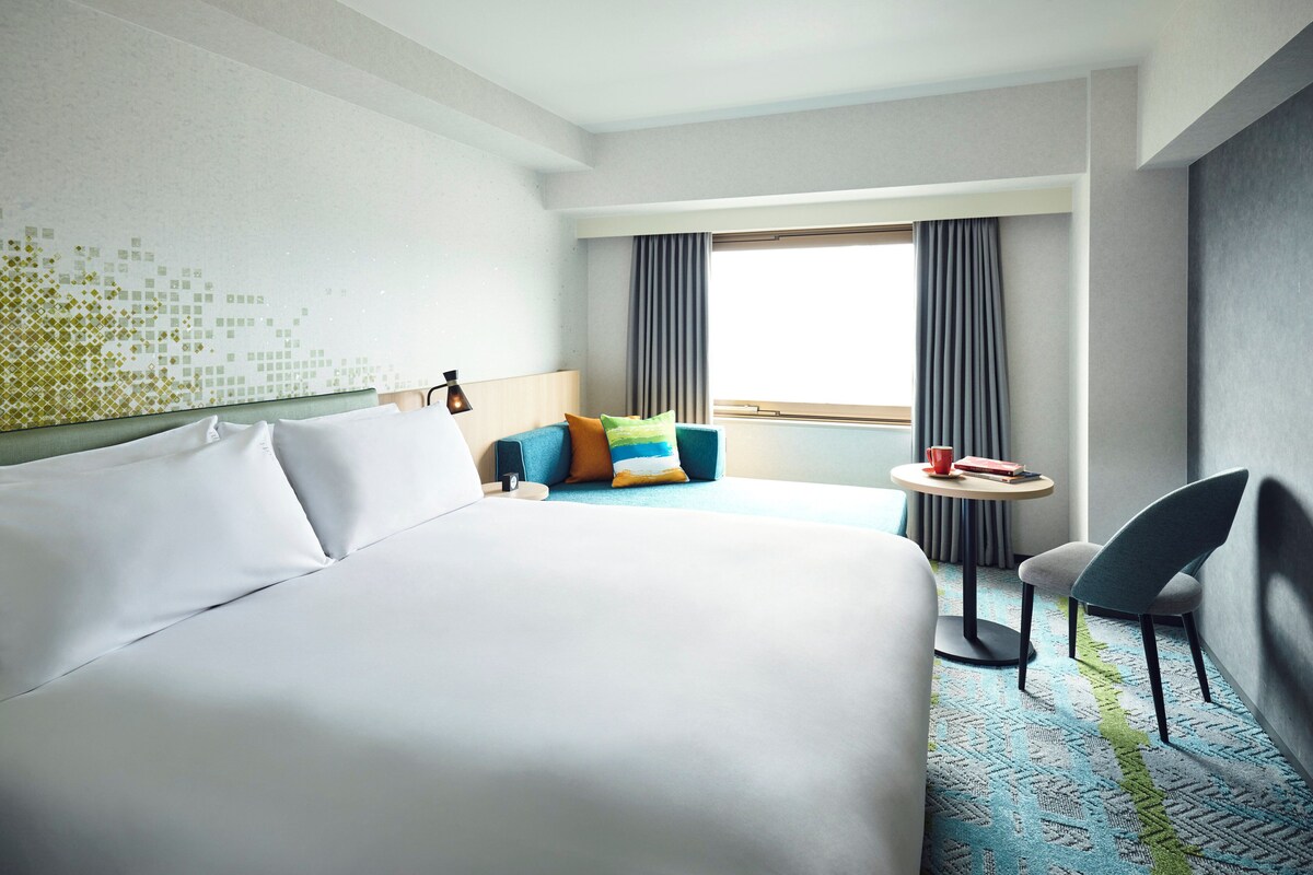 A hotel room with a large bed, a small table with a chair, a sofa with colorful cushions, and a view of the outdoors through a window.