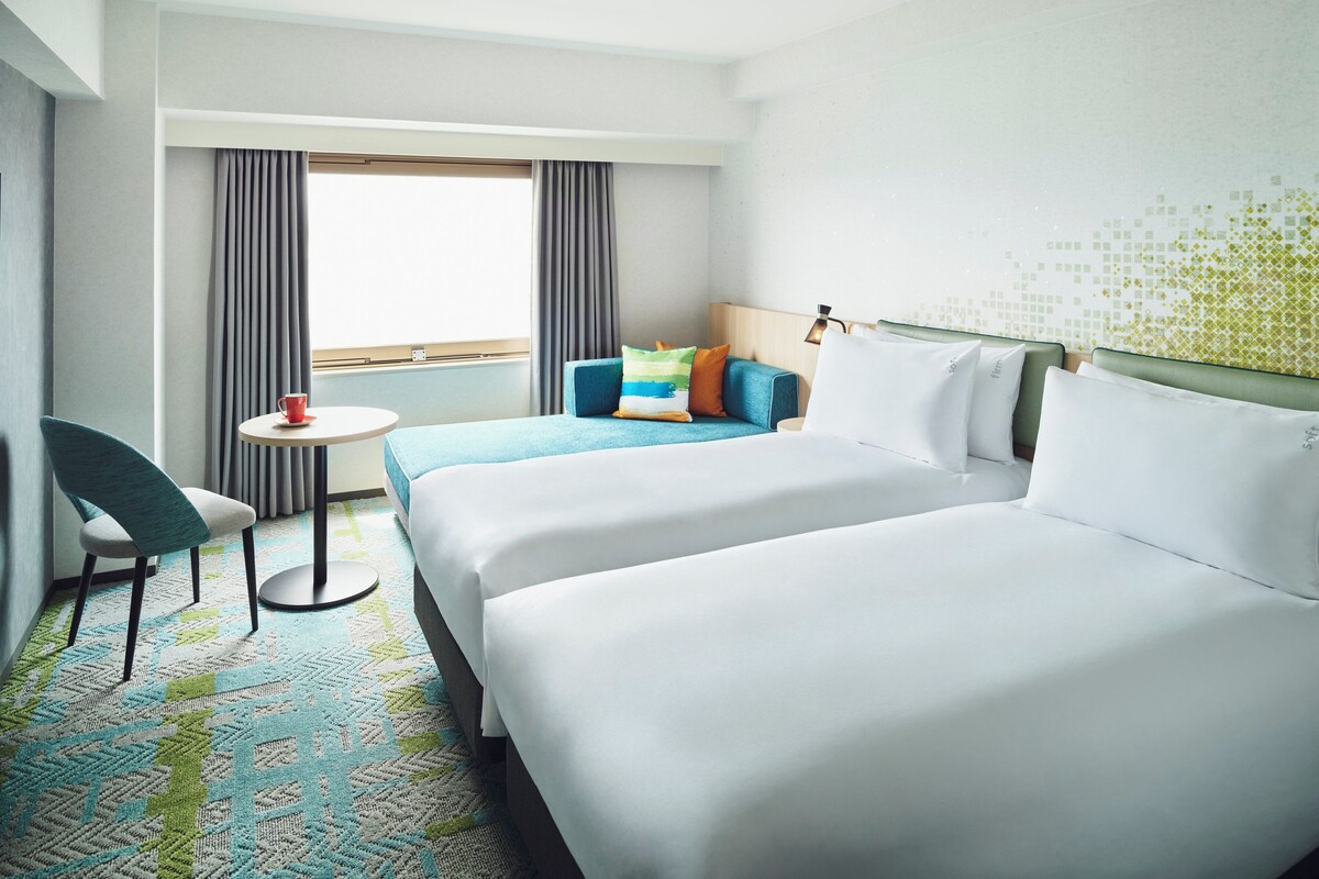 A hotel room with two beds, a small table, a chair, and a window with curtains.