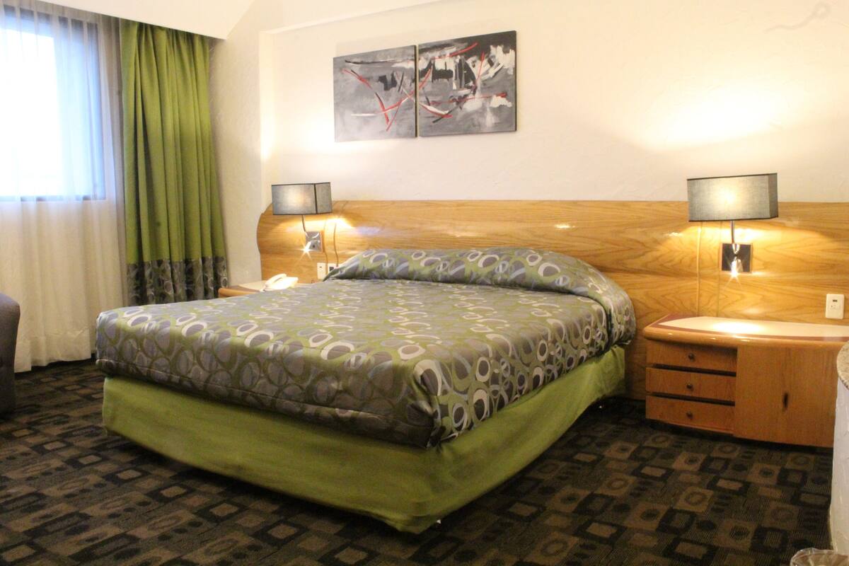 A hotel room with a bed, two bedside tables, a nightstand, a wall-mounted artwork, and a window with curtains.
