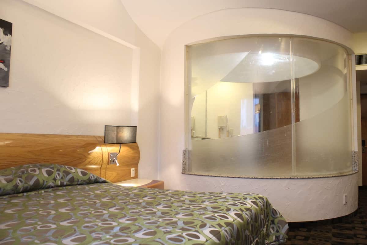 A hotel room with a curved mirror, a bed with patterned bedding, a bedside table with a lamp, and a wooden headboard.