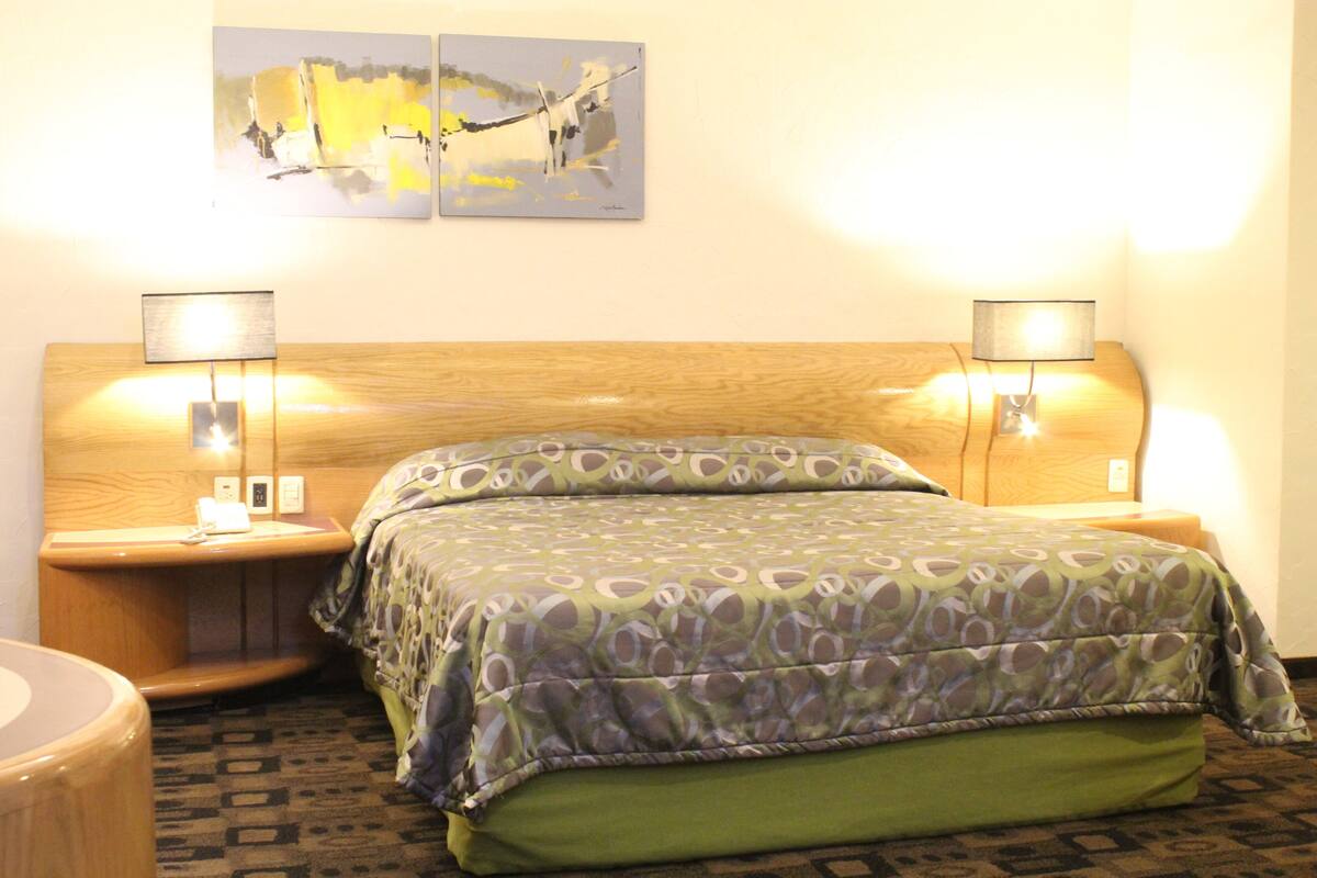 A hotel room with a wooden headboard, a green and gray patterned bedspread, two bedside lamps, and a painting above the bed.