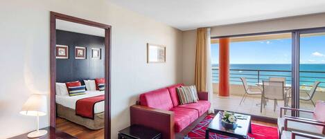 A hotel room with a bed, a red sofa, a small table, and a balcony with a view of the ocean.