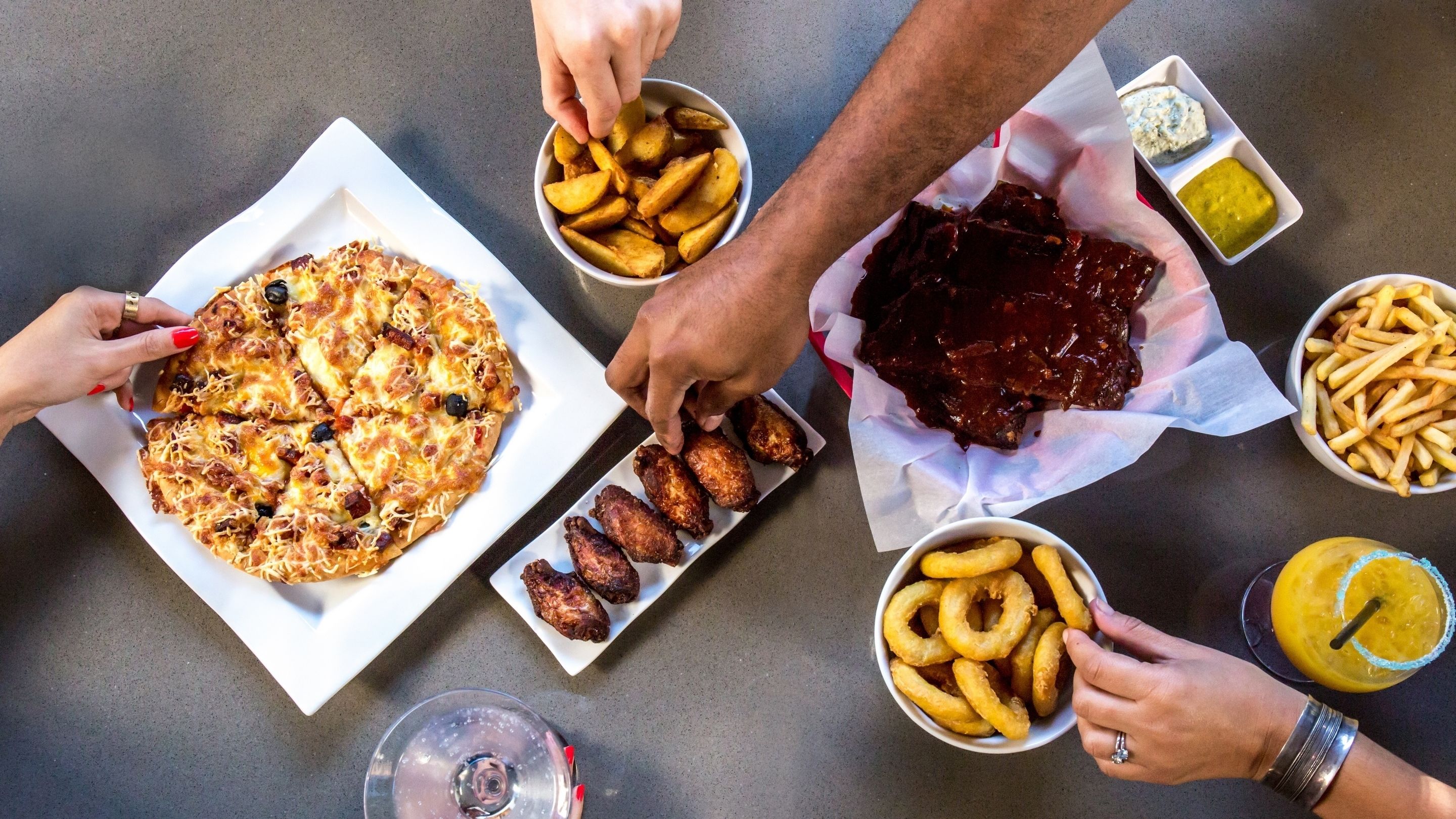 Et bord med forskellige spiser, herunder pizza, kyllingevinger, pommes frites, løgringe og en drik.