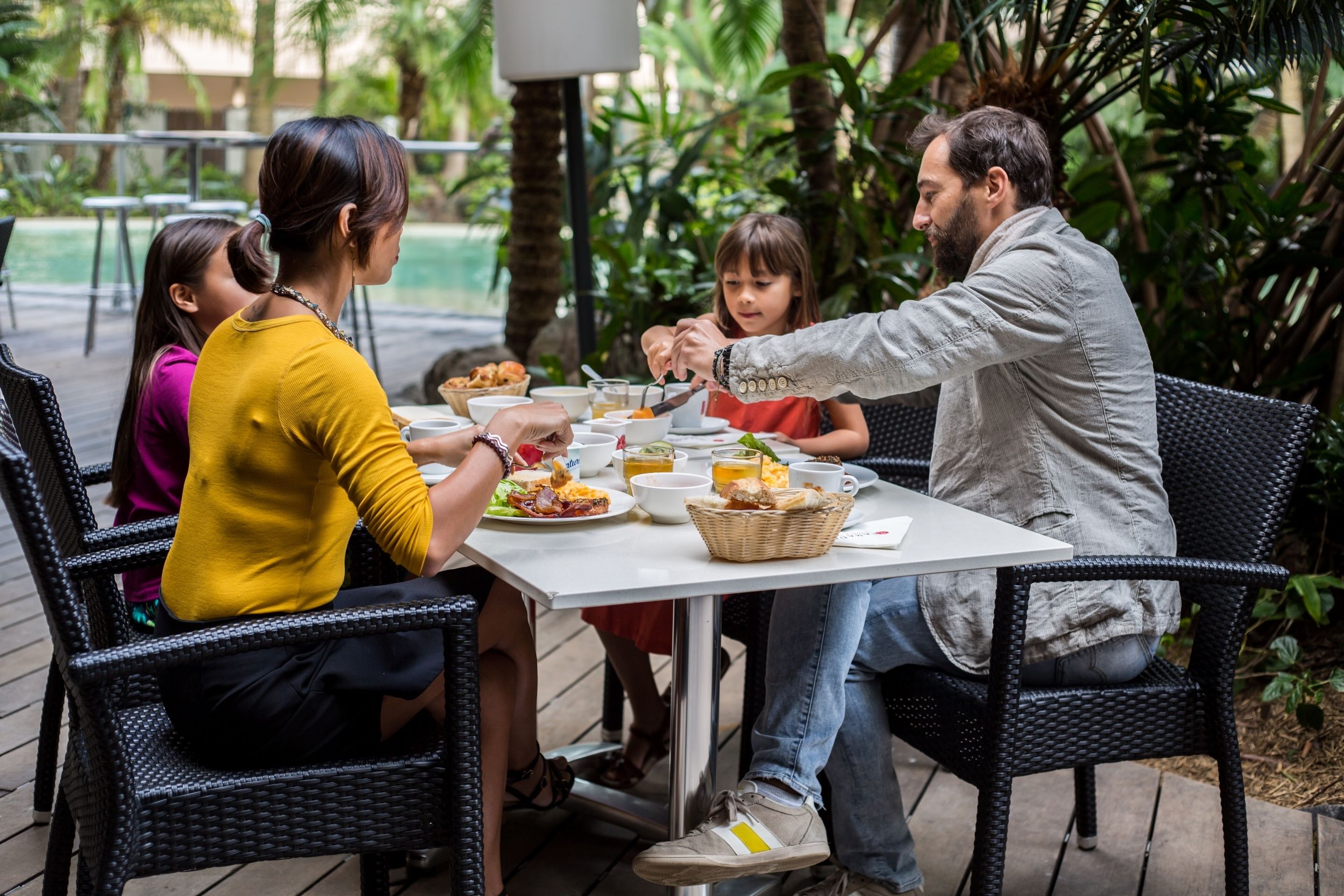 A group of people dining at an outdoor table with a white tablecloth, surrounded by greenery.