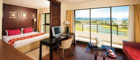 Habitación de hotel con cama, escritorio y un amplio ventanal que ofrece vistas a un campo de golf y una masa de agua.