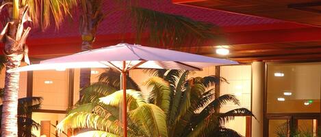Un espace de restauration en plein air, avec des tables et des chaises, entouré de palmiers et d’une piscine.