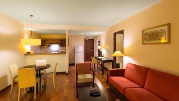 Habitación de hotel con comedor, sofá rojo, escritorio con lámpara y cocina al fondo.