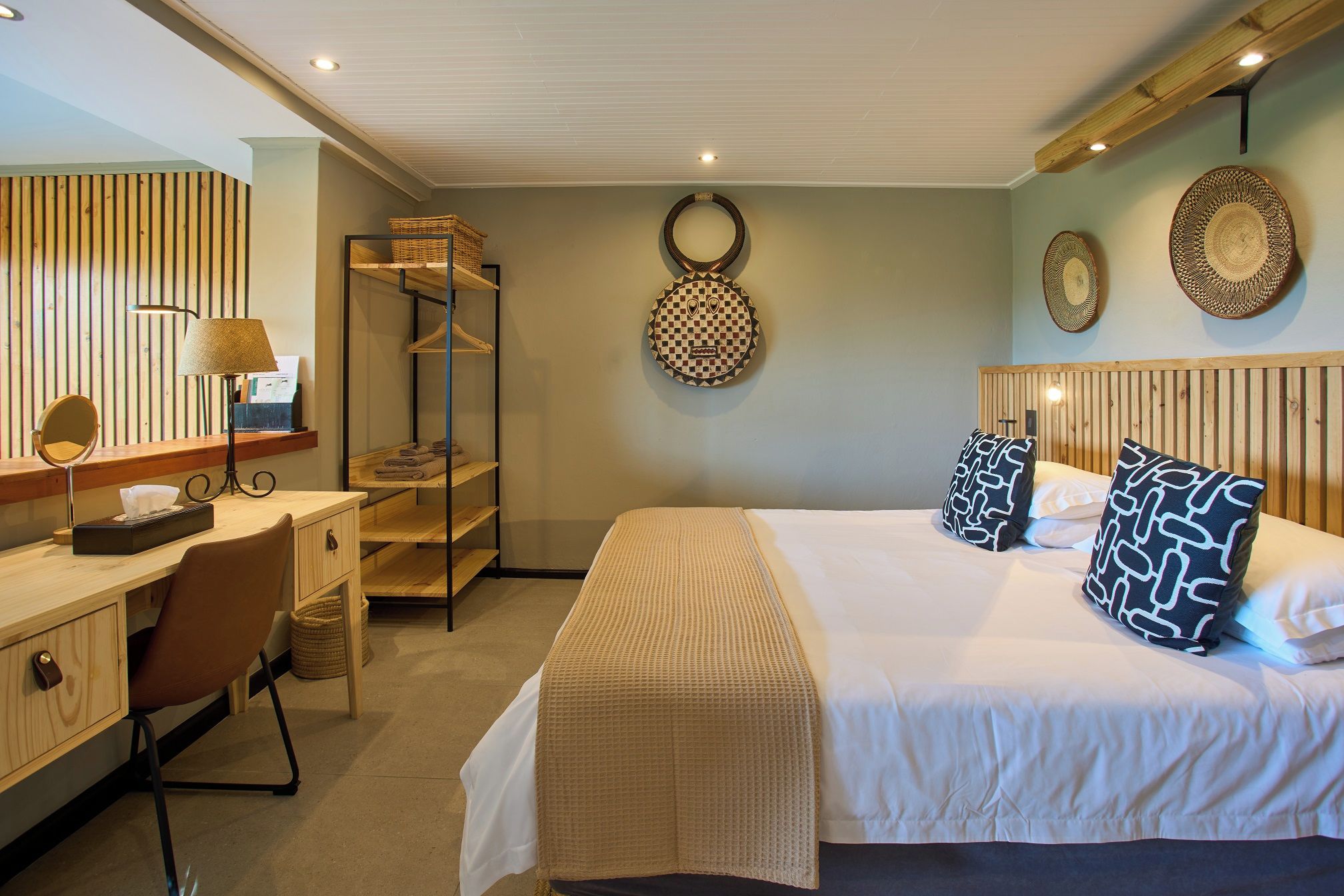 A hotel room with a bed, a desk with a lamp, a mirror, and a shelf with towels.