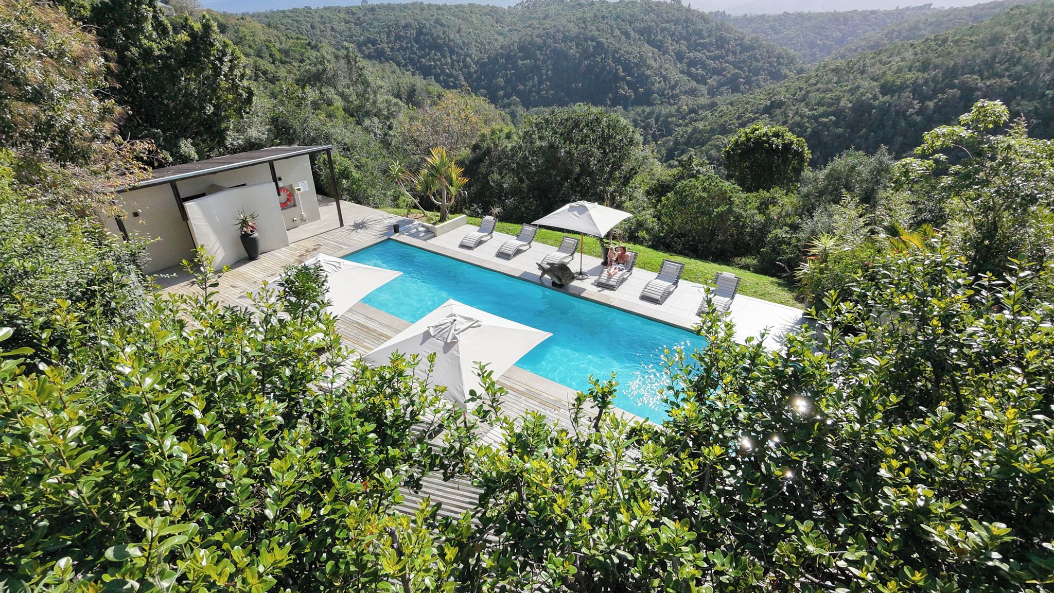 A rectangular swimming pool with a deck, surrounded by lush greenery and hills in the background.