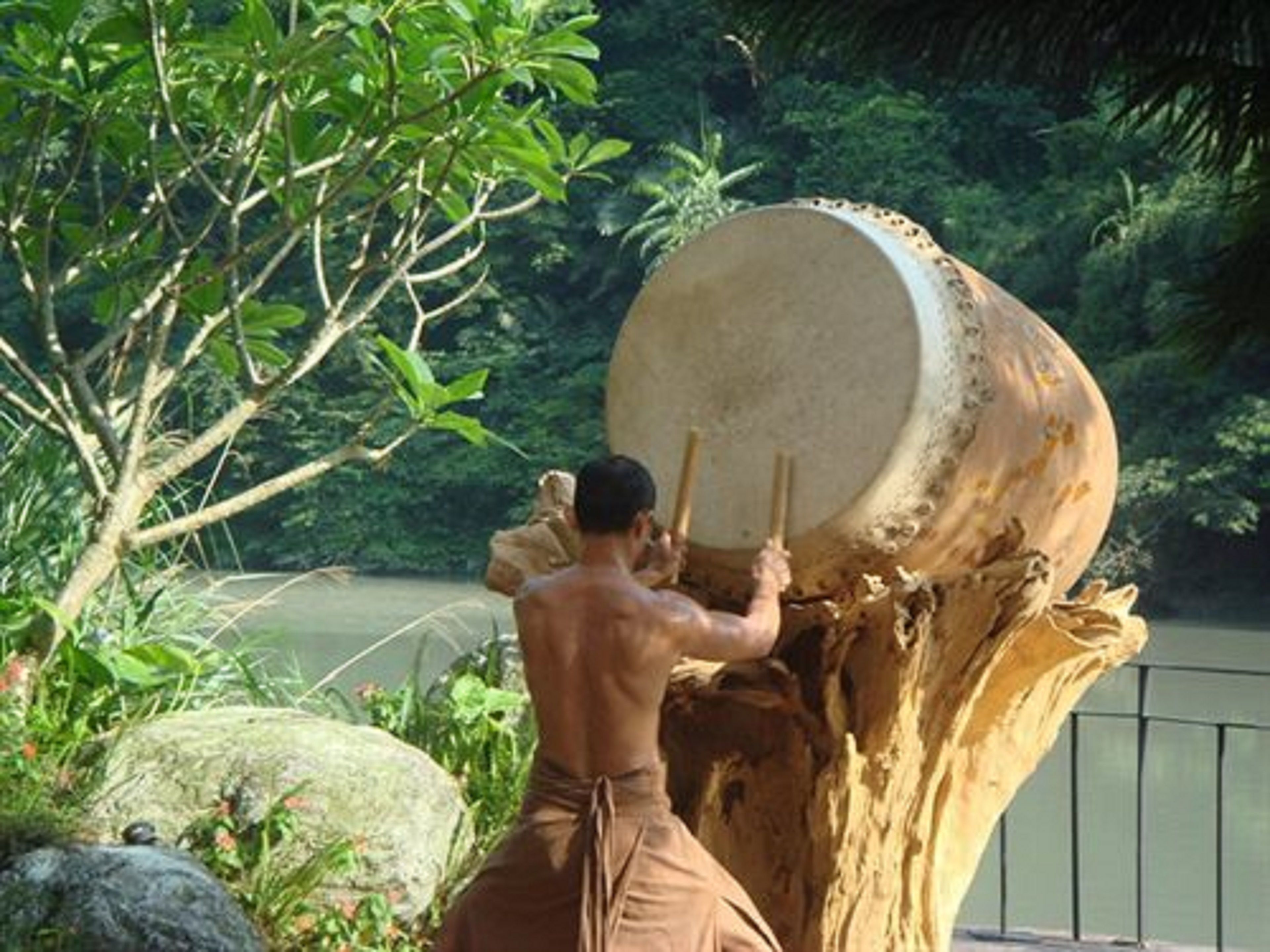 Un homme joue d’un grand tambour à l’extérieur, avec des arbres et une étendue d’eau en arrière-plan.
