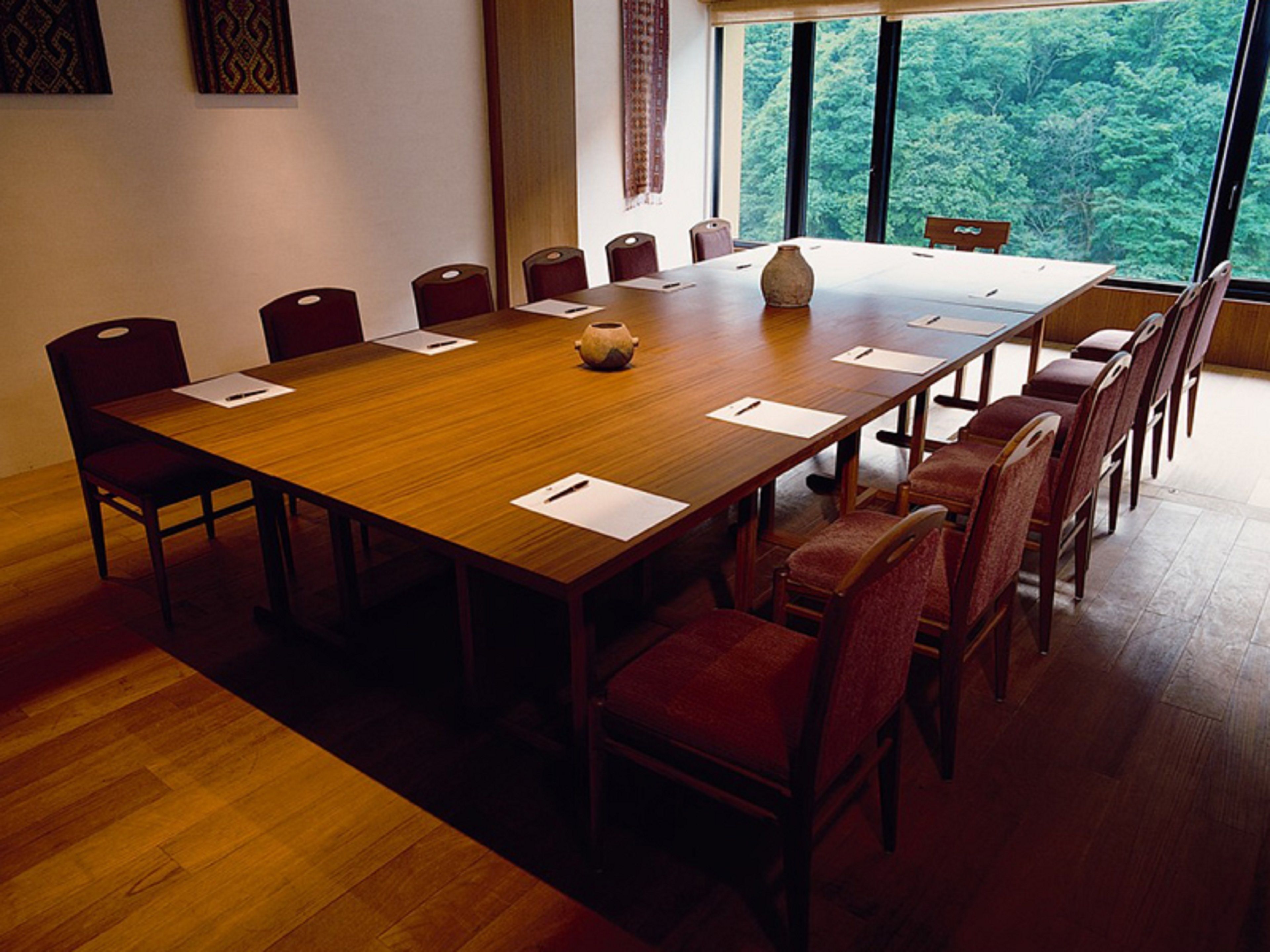 A conference room with a large wooden table, red upholstered chairs, and a view of a forest through large windows.