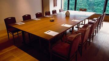 A conference room with a large wooden table, red upholstered chairs, and a view of a forest through large windows.