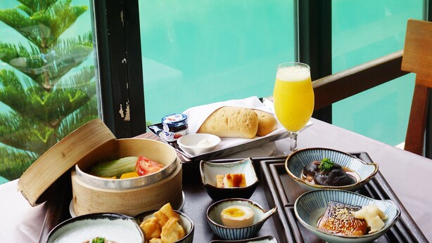 A tray of breakfast with various dishes and a glass of orange juice, viewed through a window overlooking a turquoise lake and lush greenery. - Volando Urai Spring Spa and Resort (New Taipei City)