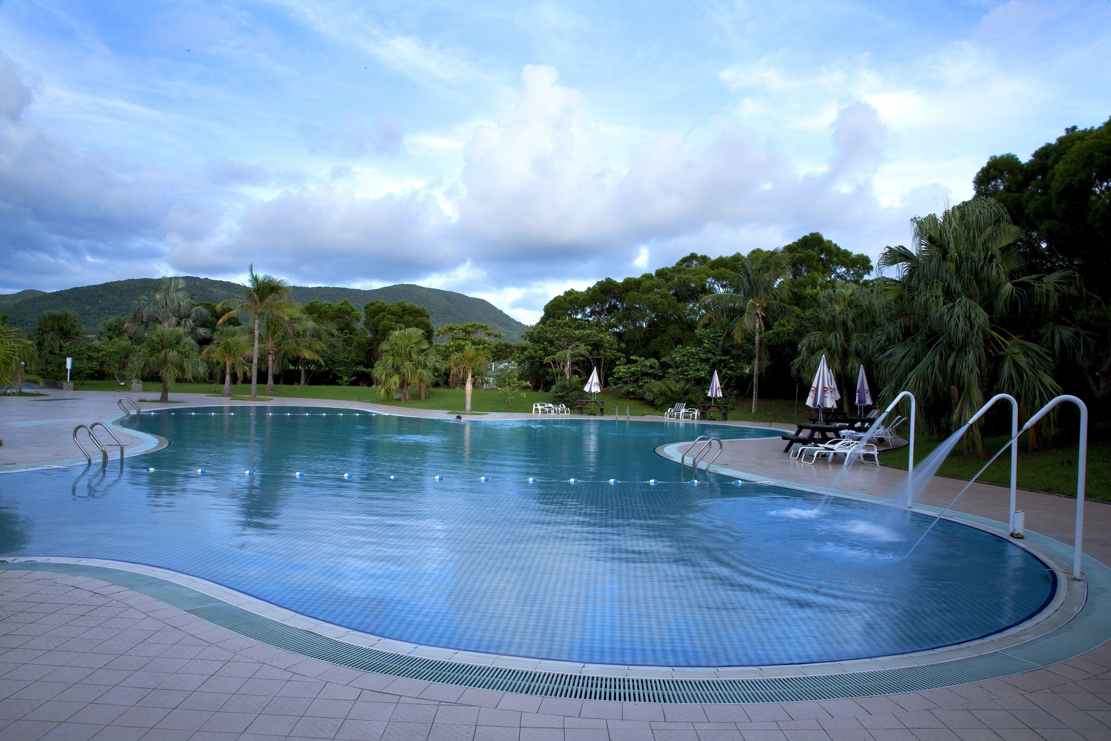 Una piscina con plataforma de baldosas, rodeada de palmeras y un paisaje montañoso.