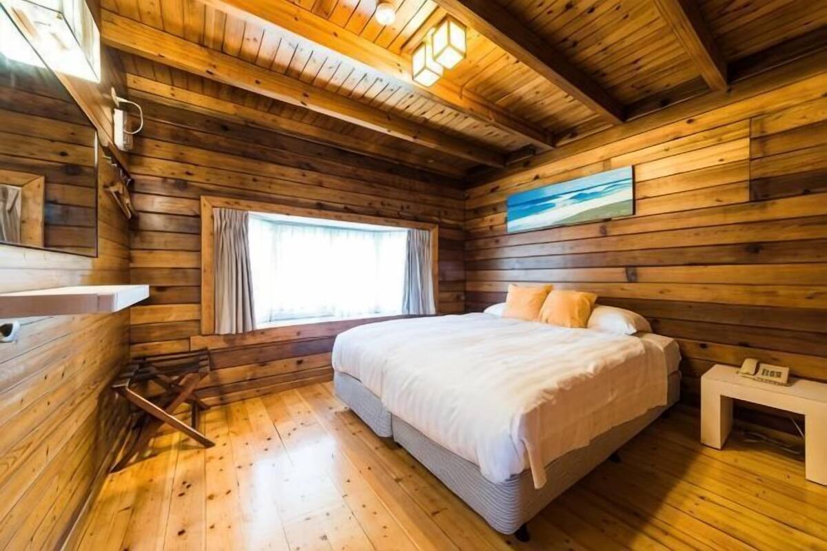 A wooden cabin room with a bed, a window with curtains, a painting on the wall, and a small bedside table.