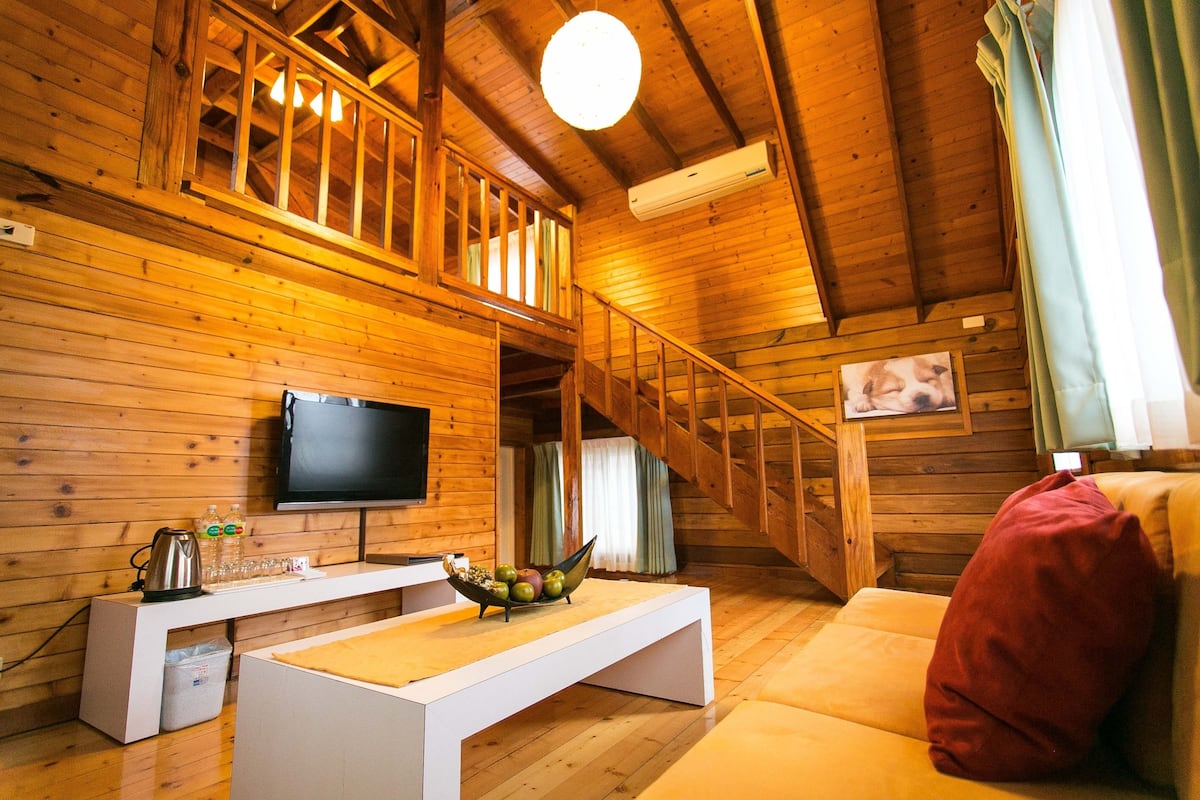 A wooden cabin interior with a TV, white furniture, a staircase, and a red pillow.