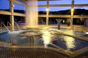 A heated outdoor pool with multiple water features, including a central fountain and surrounding hot tubs, surrounded by a covered area with seating and potted plants. - Kentington Resort (Manzhou)