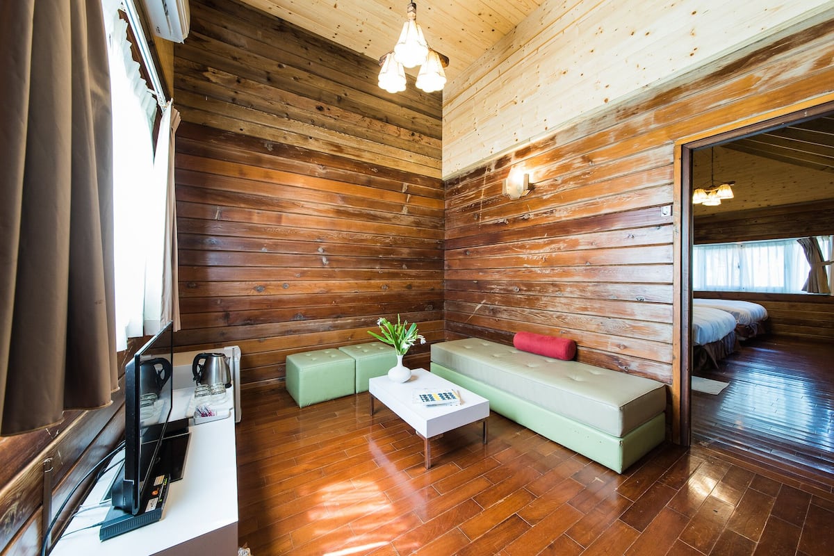A cozy cabin-style room with wooden walls, a wooden floor, a small table with a vase, a green sofa, and a bed in the adjacent room.