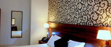 A hotel room with a large bed, a wooden headboard, a bedside table with a lamp, a telephone, and a mirror on the wall.