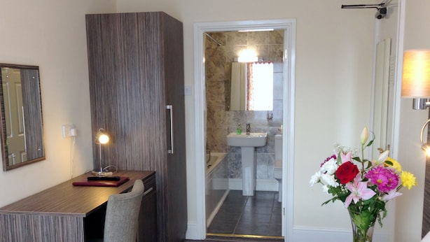 A hotel room with a wooden desk, a bed, a bathroom with a sink and mirror, and a vase with flowers on the nightstand. - Ely House Hotel (Wolverhampton)