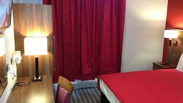 Habitación de hotel con un escritorio de madera, una silla, una cama con colcha roja, una cortina roja y dos lámparas de noche.