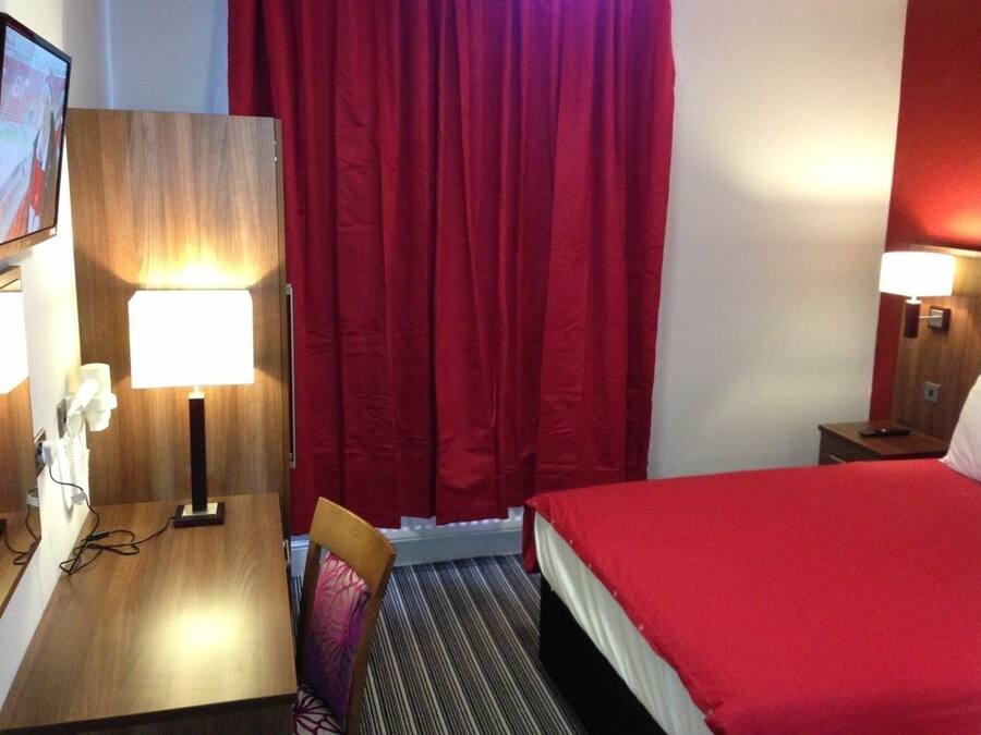 Quarto de hotel com uma secretária de madeira, uma cadeira, uma cama com colcha vermelha, cortina vermelha e duas luminárias de cabeceira.