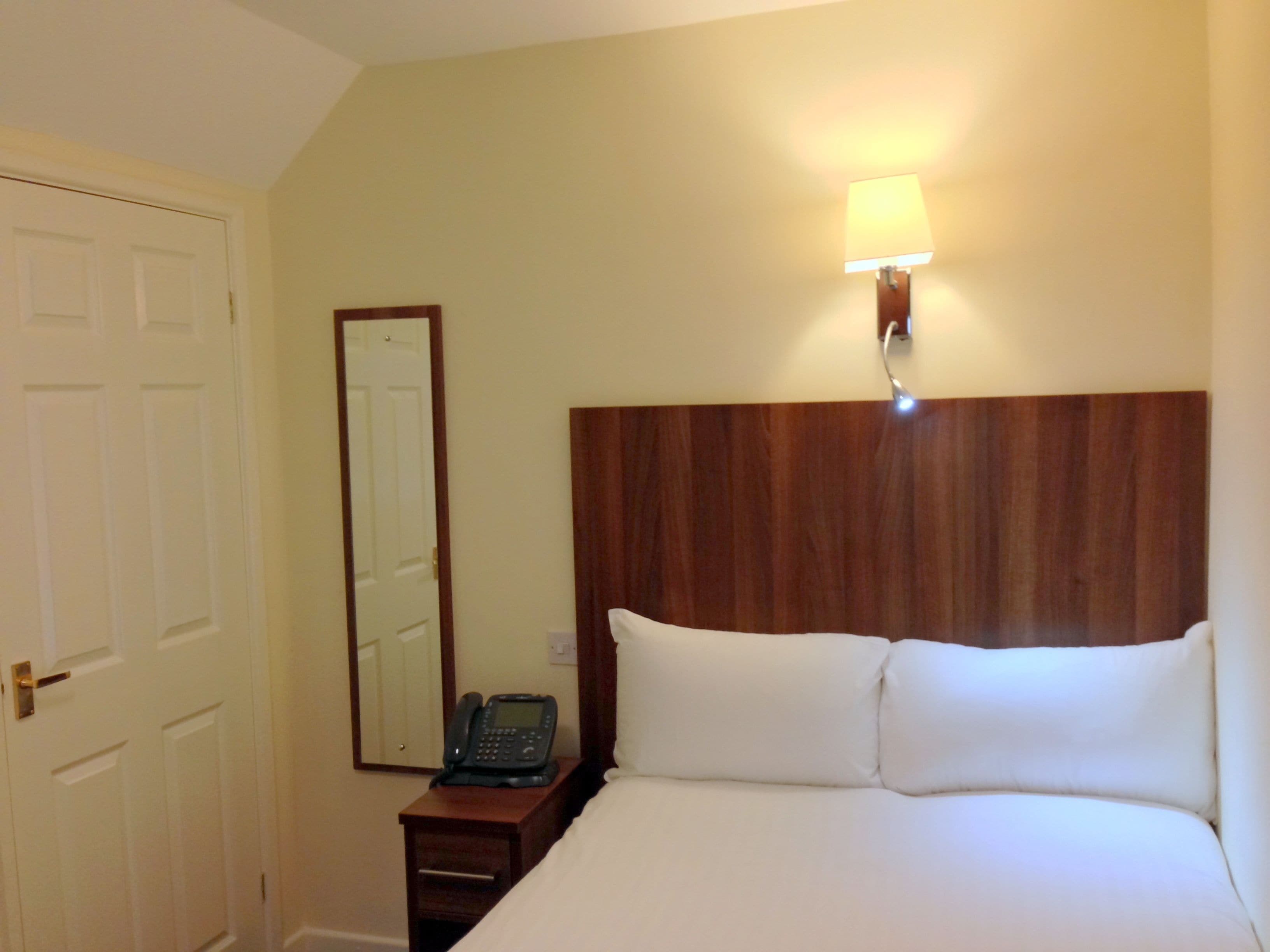 A hotel room with a wooden headboard, a bedside table with a telephone, a wall-mounted light, and a door.