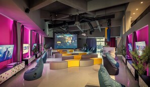A modern lounge area with multiple seating options, a large screen displaying a video game, and ambient lighting.