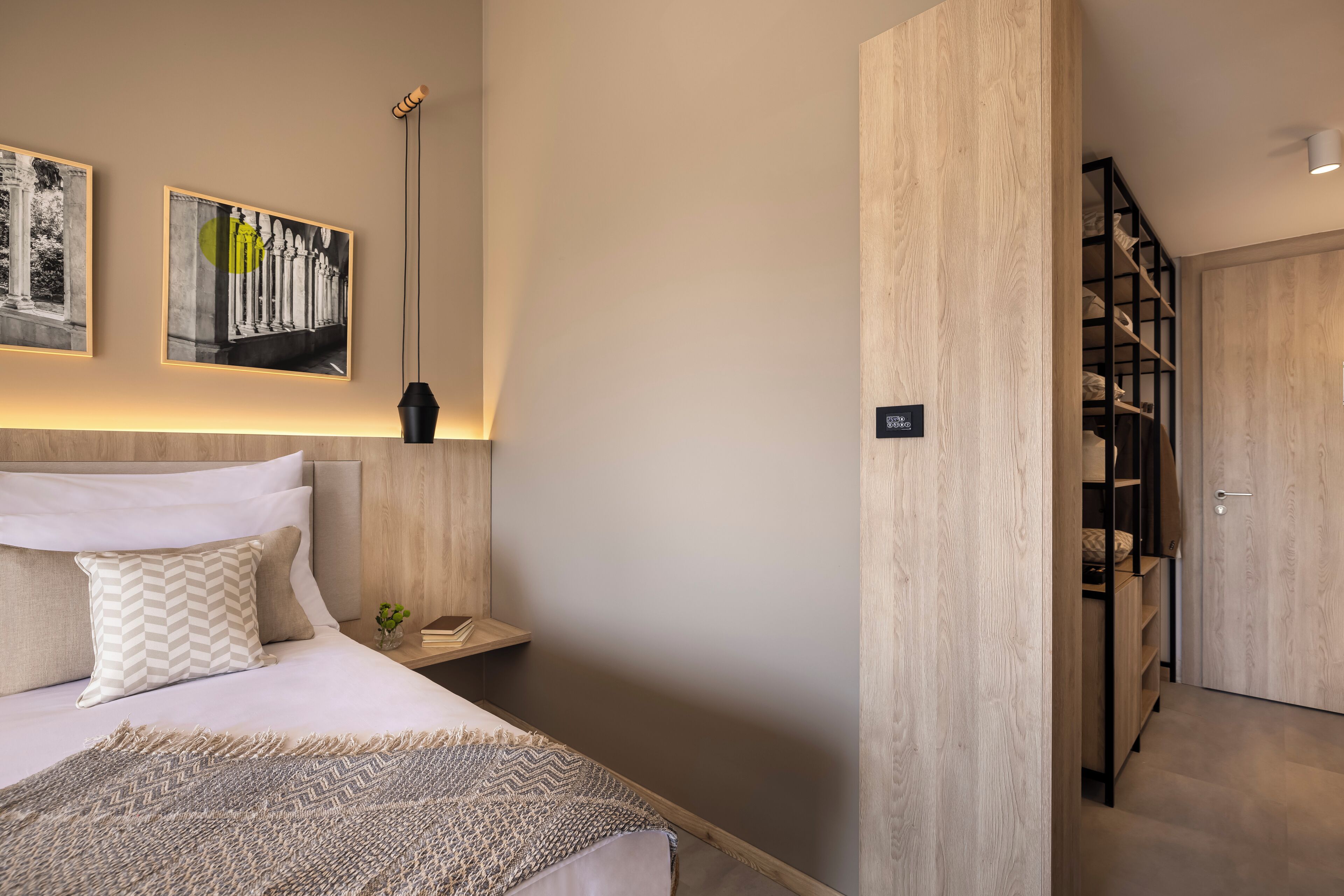 Ein modernes Hotelzimmer mit einem ordentlich bezogenen Bett, einem Nachttisch mit Lampe und einem an der Wand befestigten Regal.