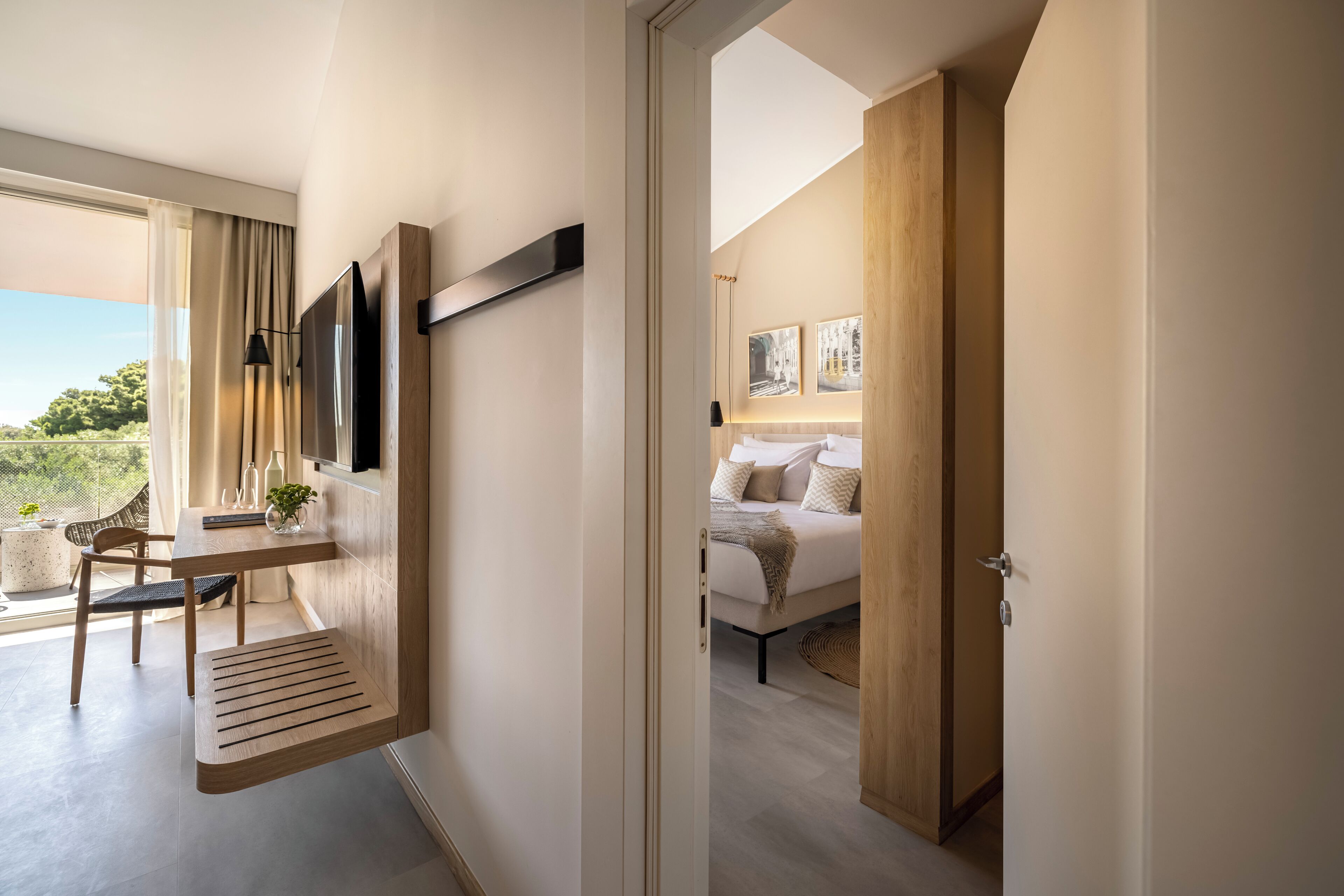 Een moderne hotelkamer met een houten bureau, een bed met wit beddengoed en een groot raam met uitzicht op groen.