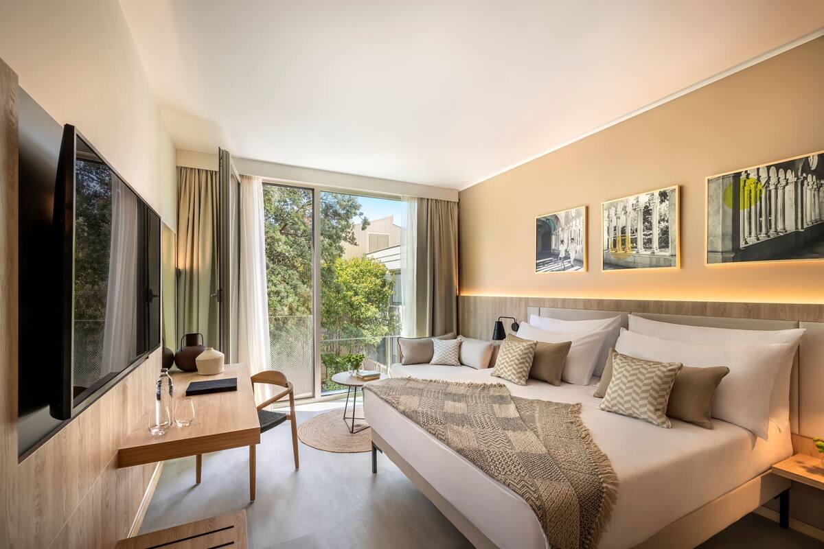 A modern hotel room with a large bed, a desk, a TV, and a balcony with a view of trees.