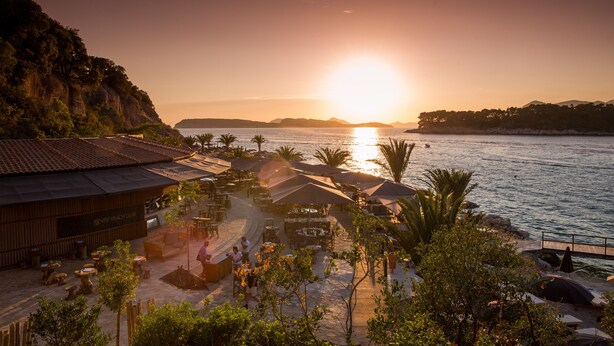 A beachfront resort with outdoor seating areas, a wooden building labeled "Ocean Beach Club," and a view of the sunset over the water. - Valamar Tirena Hotel (Dubrovnik)