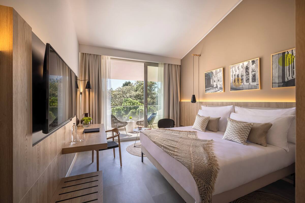 A modern hotel room with a large bed, a desk with a chair, a flat-screen TV mounted on the wall, and a balcony with a view of greenery.